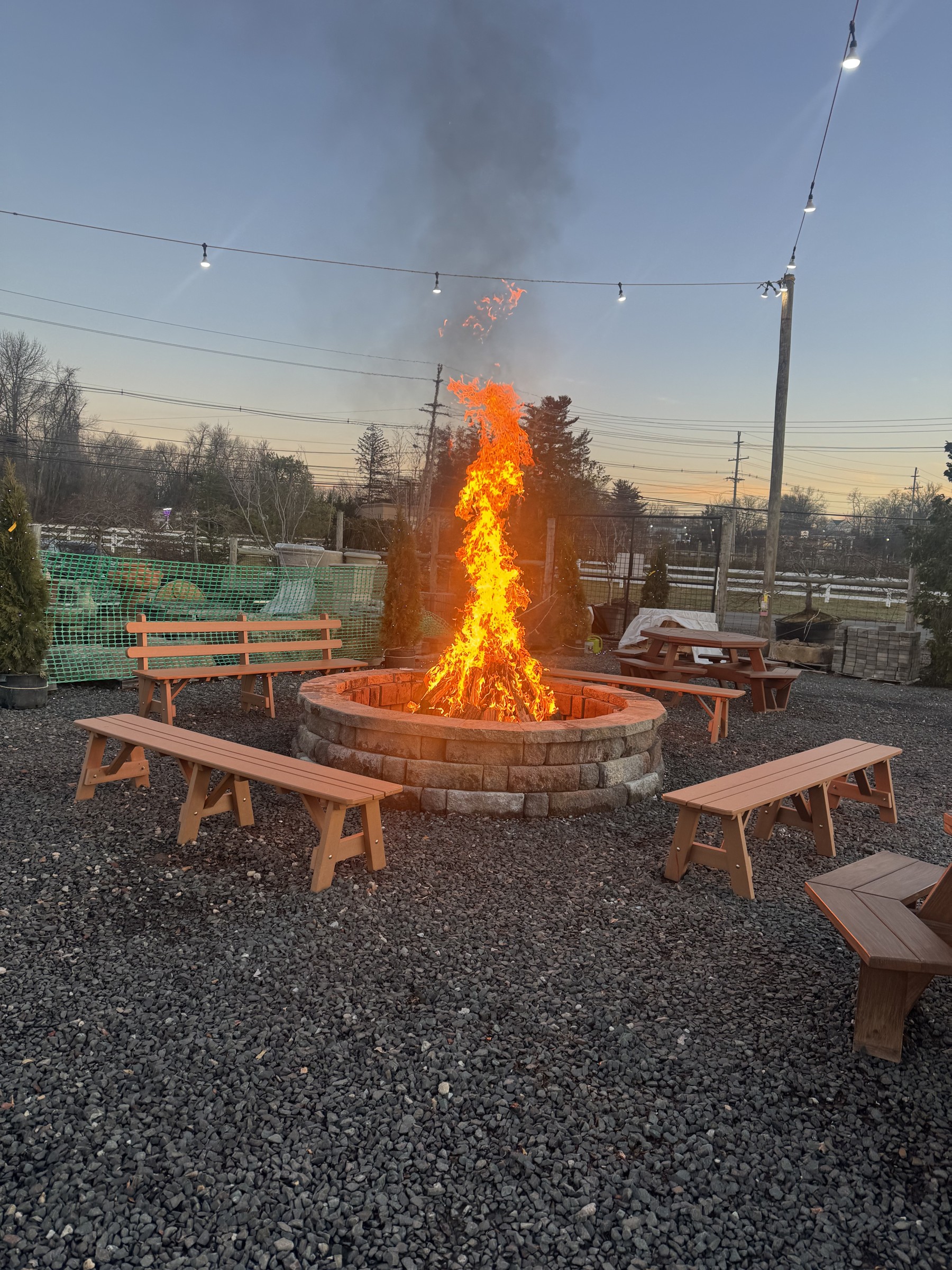 Bonfire glowing at dusk in a stone fire pit surrounded by wooden benches and string lights at Maple Leaf Farms